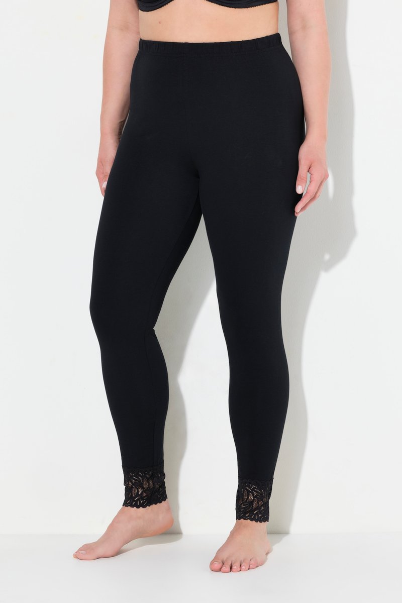 Donna che indossa leggings neri con vita elastica e rifinitura in pizzo alle caviglie, in piedi a piedi nudi su sfondo bianco.