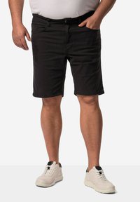 Pantalones cortos negros de tejido ligero con corte recto y pasadores para cinturón, combinados con unas zapatillas de color claro que tienen toques negros.