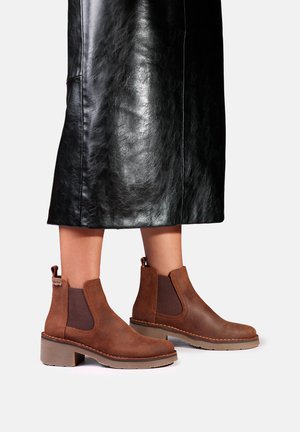 Jambes portant des bottines en cuir marron avec côtés élastiques et talons bloc, associées à une jupe en cuir noire sur un fond uni.