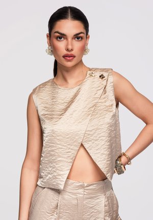 Kobieta o ciemnych włosach w beżowej, teksturowanej, bezrękawnej bluzce i pasujących spodniach, nosząca duże kolczyki i wiele bransoletek, pozująca z ręką na biodrze.