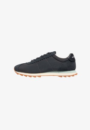 Scarpa sportiva nera che presenta una combinazione di materiali lisci e scamosciati, bordo delle lacci ondulato, suola in gomma e un bordo chiaro.