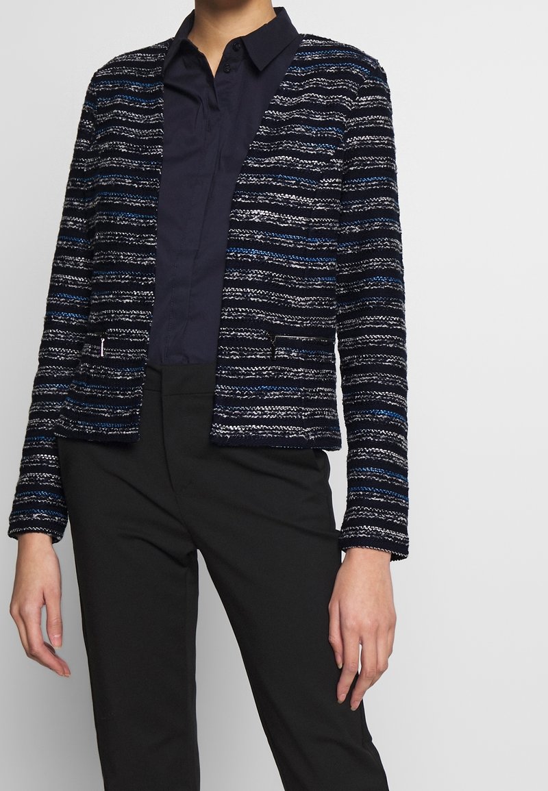 Personne portant une chemise col bleu marine, un pantalon noir et un blazer texturé rayé noir, blanc et bleu avec des poches à l'avant.