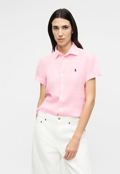 Femeie cu păr drept și închis la culoare, îmbrăcată într-o cămașă roz deschis cu mâneci scurte și pantaloni albi, stând cu o mână în buzunar.