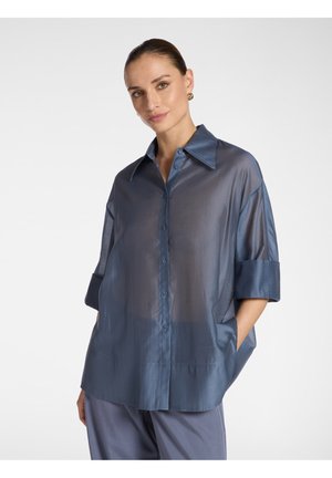 Frau trägt eine lockere, durchsichtige blaue Bluse mit Knöpfen und hochgekrempelten Ärmeln sowie passende blaue Hosen, steht mit den Händen in den Taschen.