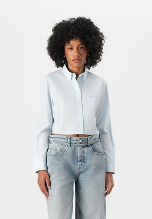 Camisa abotonada recortada de color azul claro con mangas largas y cuello camisero. Tejido texturizado con rayas verticales, con una sutil marca.