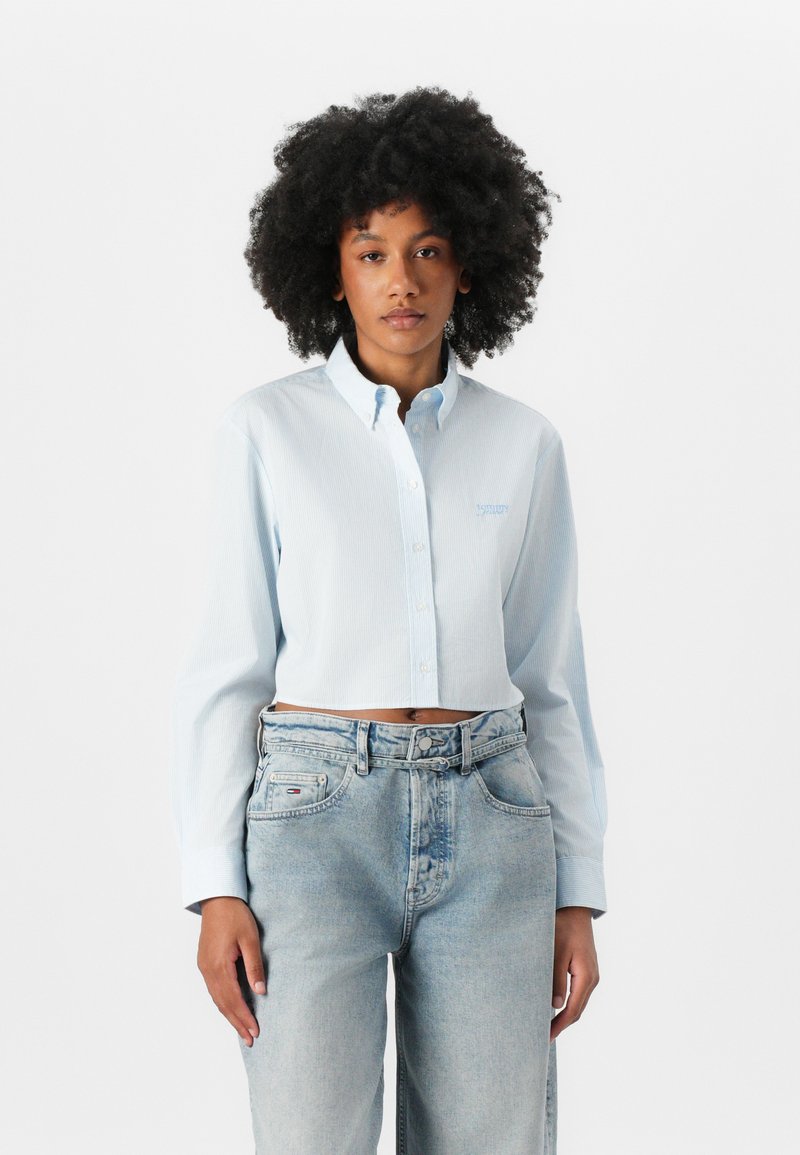 Light blue cropped button-up shirt with long sleeves and a collared neck. Textured fabric with vertical stripes, featuring subtle branding.