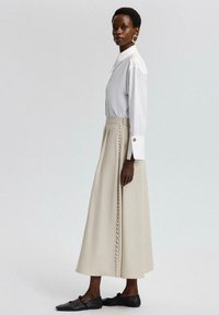 Witte bloes met een kraag, gecombineerd met een beige rok met een rij knopen aan de zijkant; model draagt zwarte flats en grote oorbellen.