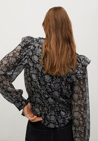 Blusa nera con motivo floreale, maniche lunghe trasparenti, dettagli arricciati sulle spalle e vita aderente, vista da dietro.