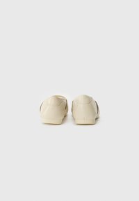 Beige slip-on sko med en glatt, skinnaktig finish, avrundet bak, og et minimalistisk design, uten synlig maskinvare eller mønstre.