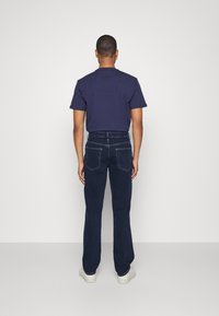 Blå jeans med en avslappnad passform, med två bakfickor och sömnadsdetaljer, kombinerade med en mörk t-shirt. Vita sneakers syns längst ner.