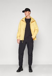 Gul vattentät jacka med huva, som är kombinerad med en svart t-shirt och svarta cargobyxor. Svarta snörkängor. Enkel design och praktisk passform.
