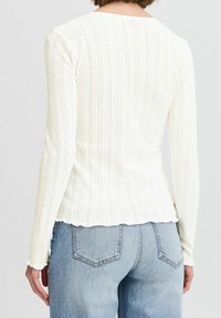 Biała bluzka z długim rękawem, prążkowana, z falbaniastym brzegiem, z pionowymi paskami i teksturowanym wzorem. Noszona z jasnoniebieskimi dżinsami.