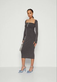 Femme portant une robe midi longue gris foncé, ajustée et décolleté, des escarpins bleus avec des nœuds, et tenant un petit sac à main argenté.