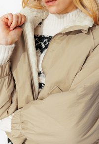 Doudoune beige avec un col doux doublé de fourrure, des manches surdimensionnées et une fermeture éclair à l'avant, portée par-dessus un pull en tricot noir et blanc.