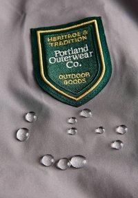 Sivi materijal s izvezenom zeleno-zlatnom oznakom "Portland Outerwear Co." i kapljicama vode na površini.