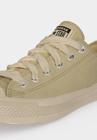Μπεζ χαμηλό sneaker Converse Chuck Taylor All Star με λευκά κορδόνια και λαστιχένια σόλα, που δείχνει το λογότυπο στη γλώσσα.