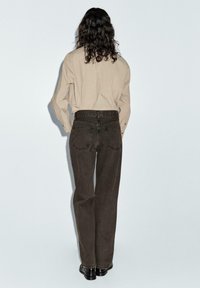 Bruna vida byxor med hög midja och fem fickor, tillsammans med en beige långärmad skjorta, sedd bakifrån. Svarta skor är på.