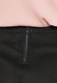 Svart fuskmockaskjol med en synlig framdragkedja och rund dragkrok, tillsammans med en ljusrosa ribbad topp.