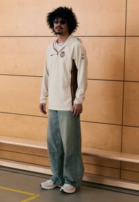 Beige Nike hoodie met bruine accenten, NBA-logo en ritssluiting kraag; gecombineerd met wijde blauwe denim jeans en witte sneakers.