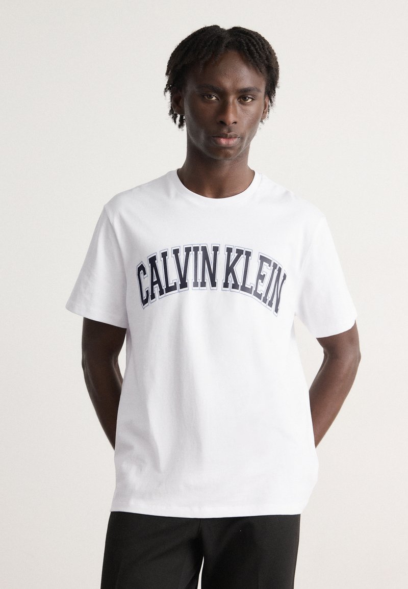 Hvit bomulls T-skjorte med en marineblå "CALVIN KLEIN"-logo i buet design over brystet, kort ermer og rund hals.