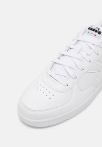 Witte leren sneaker met een gladde textuur, geperforeerd teendeel, platte veters en een logo op de tong met rode en groene accenten.