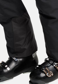 Zwarte skibotten met metalen gespen en verstelhardware, in combinatie met waterdichte zwarte skibroeken met een gladde textuur.