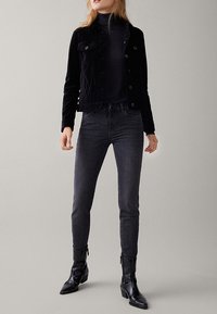 Musta pystykaarroke takki napituksella, yhdistetty tummanharmaisiin kapeisiin farkkuihin ja mustiin nahkaisiin nilkkureihin. Yksinkertainen, istuva malli, ilman näkyviä kuvioita.