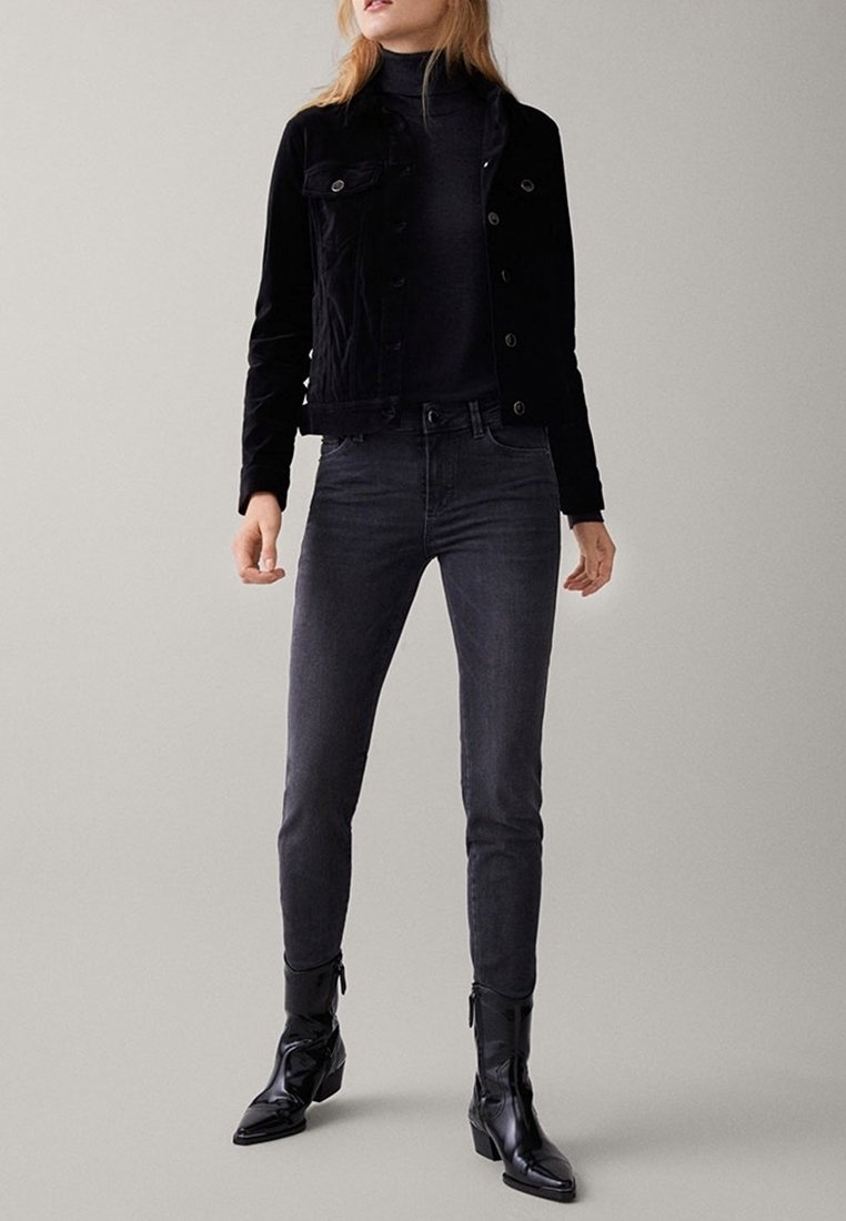 Musta pystykaarroke takki napituksella, yhdistetty tummanharmaisiin kapeisiin farkkuihin ja mustiin nahkaisiin nilkkureihin. Yksinkertainen, istuva malli, ilman näkyviä kuvioita.