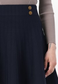 Marineblauwe geplooide rok van textuurstof, met twee decoratieve gouden knopen op de tailleband. Een hand is gedeeltelijk zichtbaar, met een tattoo.