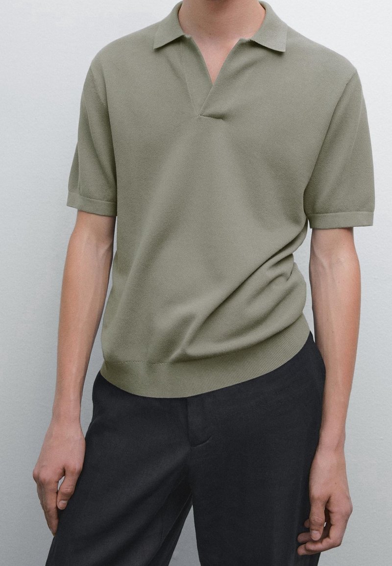 Homme portant un polo vert olive à manches courtes, rentré dans un pantalon noir, sur un fond clair uni.