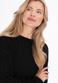 Zwarte gebreide trui met een gestructureerd patroon, voorzien van een ronde hals en lange mouwen, gedragen door een persoon met lang, golvend haar.