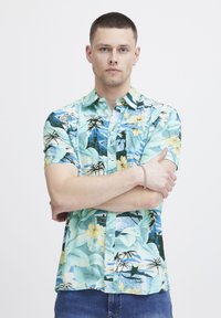 Camicetta a maniche corte, con bottoni, realizzata in un tessuto leggero di colore verde menta, caratterizzata da motivi floreali tropicali e palme con onde oceaniche blu.
