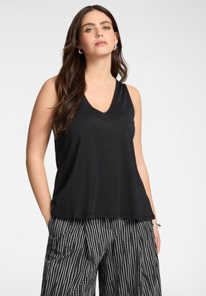 Femme aux longs cheveux bruns portant un haut noir sans manches à col en V et un pantalon large rayé noir et blanc, debout les mains dans les poches.