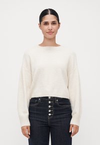 Kremfarget cropped genser med lange ermer, ribbestrikket hem og rund hals, parret med høyt liv mørk denim jeans med sølvknapper.