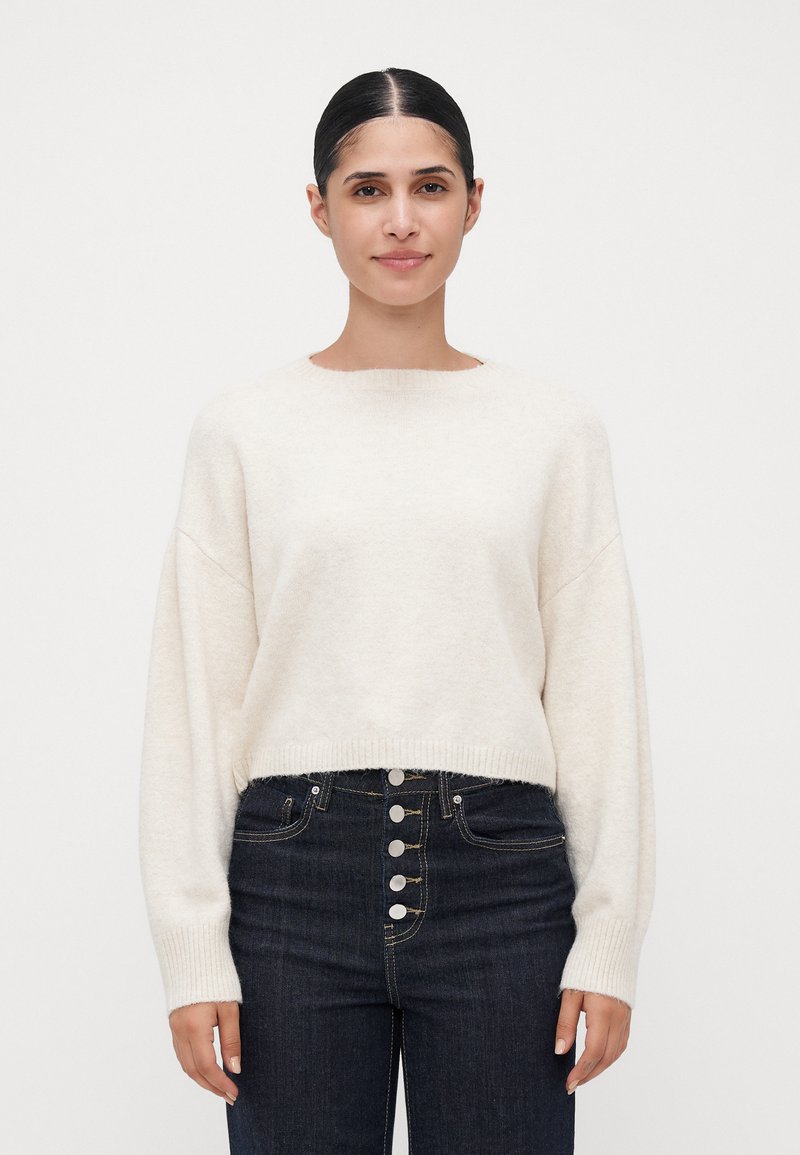 Kremfarget cropped genser med lange ermer, ribbestrikket hem og rund hals, parret med høyt liv mørk denim jeans med sølvknapper.