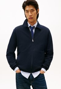 Jeune homme portant une veste zippée bleu marine par-dessus une chemise rayée bleue et une cravate à pois, les mains dans les poches et une expression neutre.