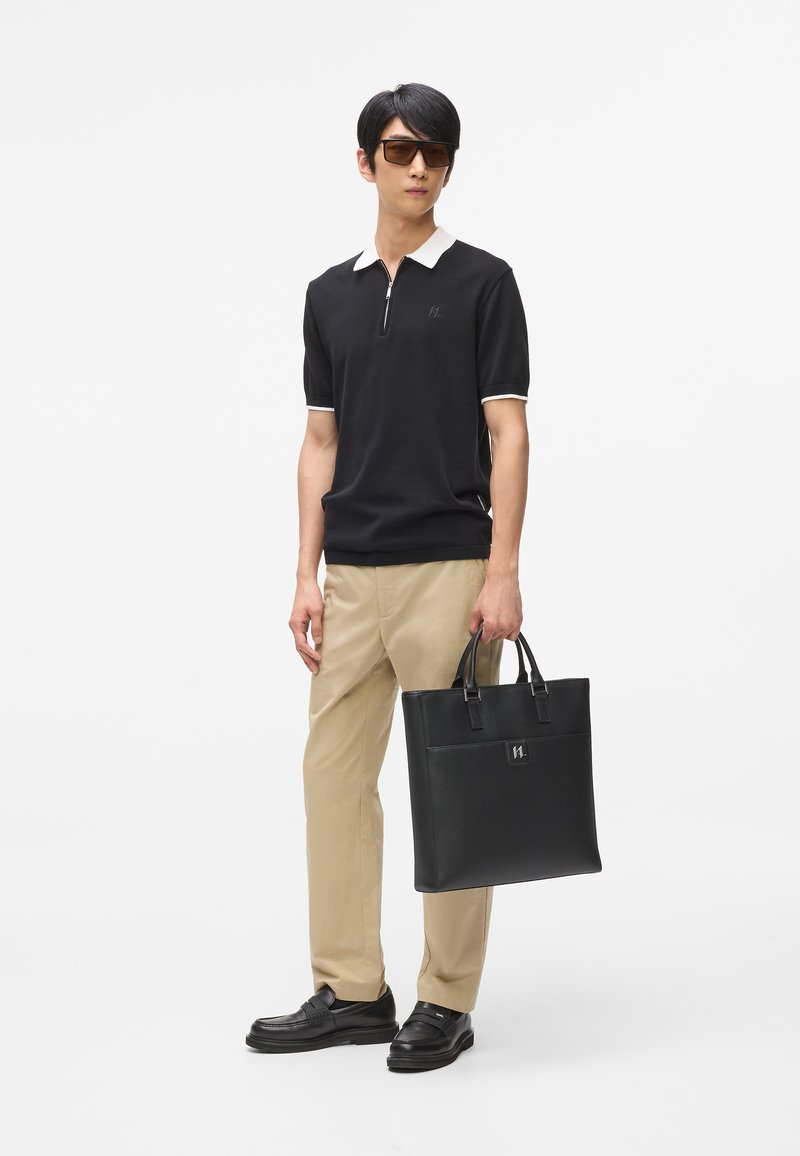 Fekete póló fehér gallérral, khaki nadrág és fekete bőr cipő. Egy fekete tote táskát tart, amely kontrasztos logó részlettel rendelkezik.