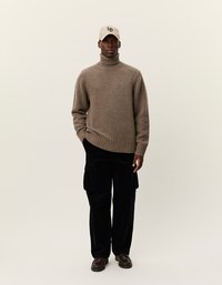 Καφέ πλεχτός πουλόβερ με ψηλό γιακά, μαύρα cargo παντελόνια, μπεζ καπέλο μπέιζμπολ με λογότυπο και καφέ παπούτσια; στέκεται σε ουδέτερο φόντο.