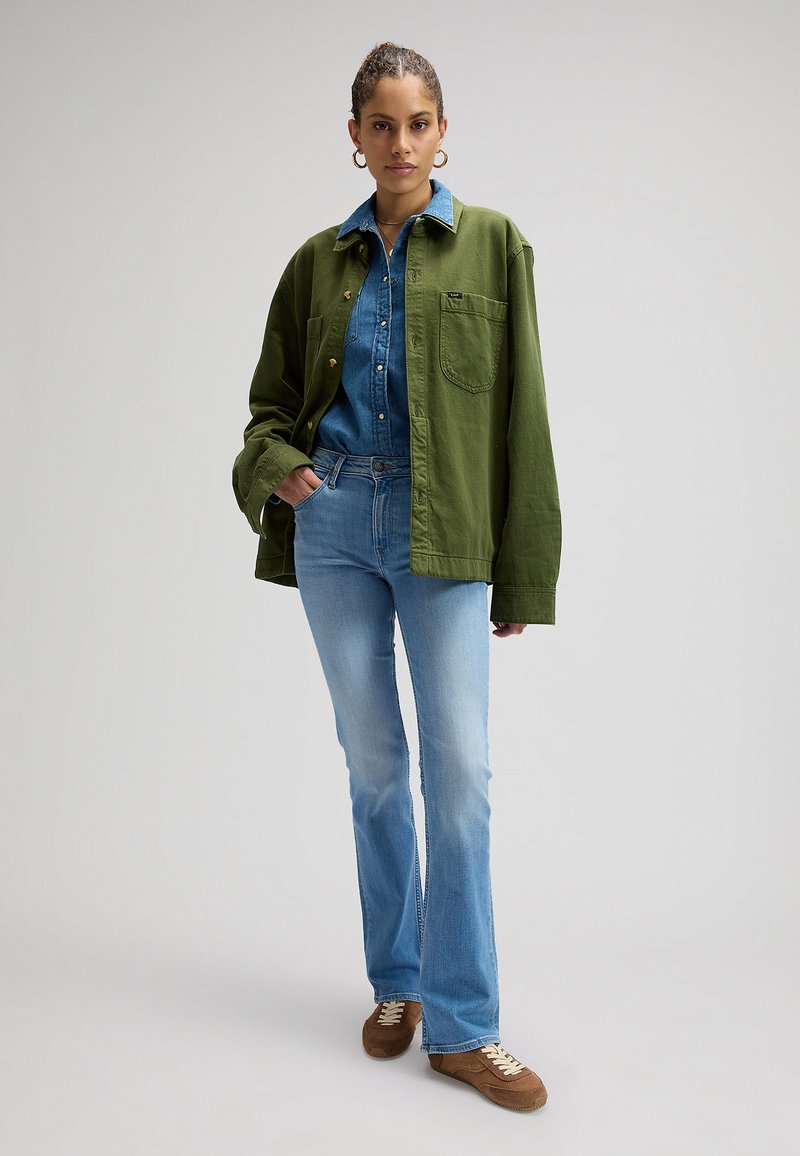 Kvinde med flettet hår iført øreringe i ringe, grøn knap-op overshirt over en denimskjorte, lyseblå jeans og brune sneakers, står oprejst.