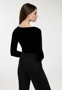 Młoda kobieta z długimi brązowymi włosami, ubrana w czarną aksamitną bluzkę z długim rękawem i czarne dżinsy z wysokim stanem, stojąca bokiem na jednolitym tle.