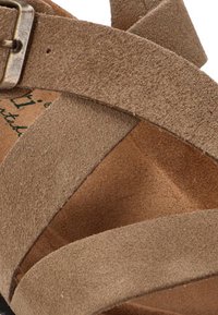 Correas de sandalia de ante color tostado cruzando sobre la plantilla, con hebilla metálica visible y parte de la suela de corcho en la parte inferior.