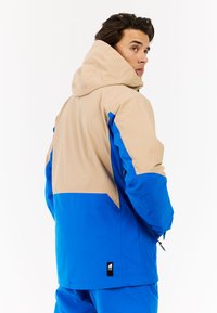 Spalvų blokais dėvima striukė, su mėlyna apatine dalimi ir smėlio spalvos viršutine dalimi, su gobtuvu, vandeniui atsparia medžiaga ir šoninėmis zip kišenėmis.