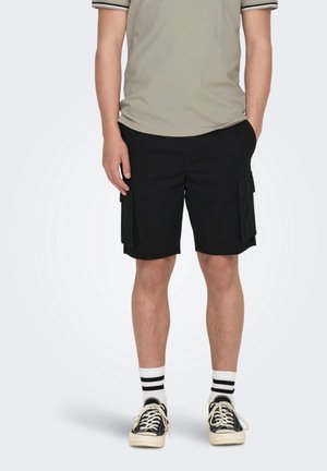 Person trägt schwarze Cargo-Shorts, beige kurzärmeliges Hemd, schwarz-weiß gestreifte Socken und schwarze Sneakers mit Schnürsenkeln.