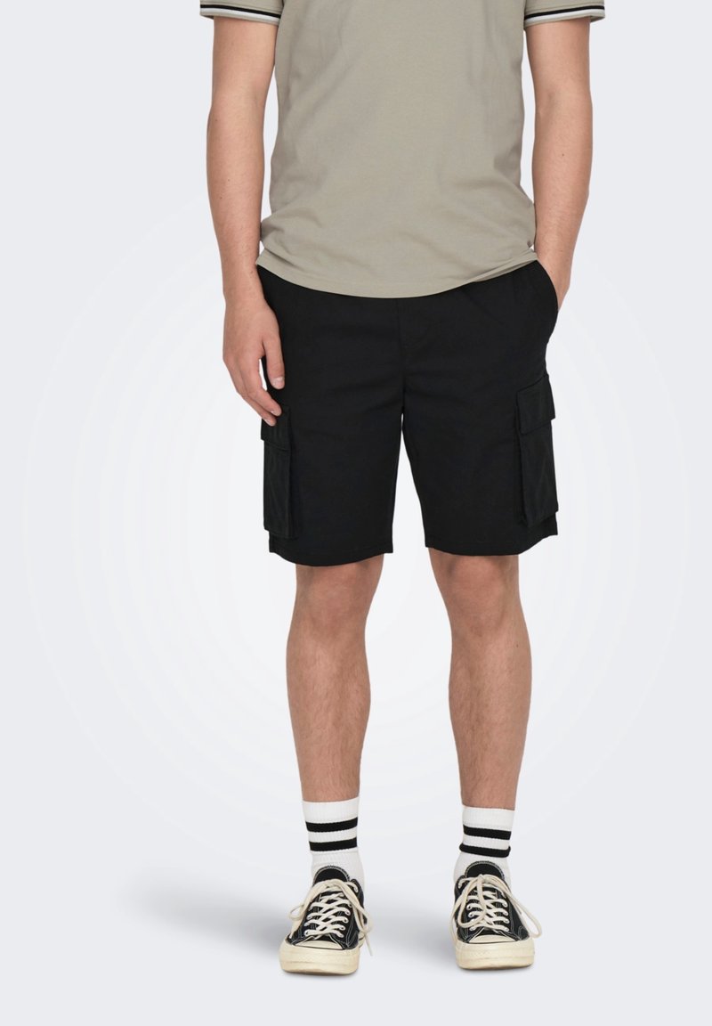 Personne portant un short cargo noir, une chemise beige à manches courtes, des chaussettes rayées noires et blanches, et des baskets noires à lacets.