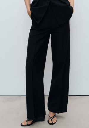 Black, wide-leg trousers with a smooth fabric, featuring a high waist and front pockets, paired with black flip-flops.