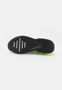 Suola di scarpa sportiva nera con superficie testurizzata, inclusi pattern in gomma per grip e tre accenti argentati. Dettaglio verde sul tallone.