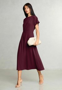 Vīnogu krāsas maksi kleita ar mezglotu vidukļa detaļu, maigu audumu, īsām piedurknēm un plūstošu svārku, kopā ar bēša krāsas krokotu rokassomiņu.