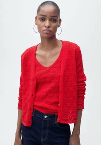 Rode gebreide cardigan met een gegolfd patroon, met daaronder een v-hals top, beiden met een gestructureerde afwerking, gecombineerd met donkere jeans.
