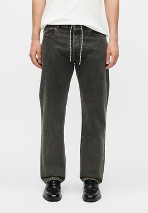 Hombre con vaqueros negros holgados de mezclilla con cintura de cordón blanco y zapatos de cuero negros, de pie frente a un fondo liso.