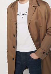 Brun trenchcoat med en slät yta, knappstängning och två framfickor över en vit t-shirt med svart text. Bärs med blåjeans.